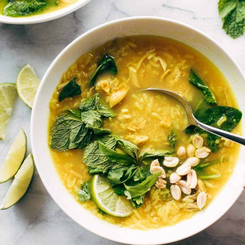 Chicken and rice soup in a bowl with a spoon. There are fresh herbs on top and lime wedges on the side.